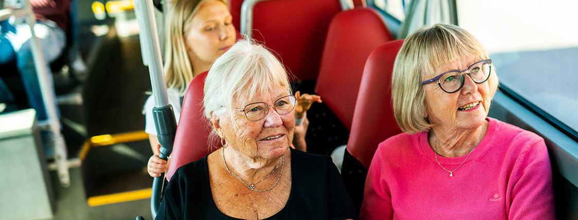 Två äldre damer sitter ombord på en buss. Bakom sitter en kvinna med ett barn.