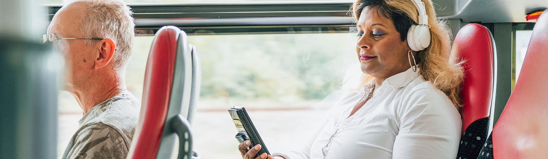 En kvinna sitter ombord på en buss med hörlurar på sig och tittar i mobiltelefonen.