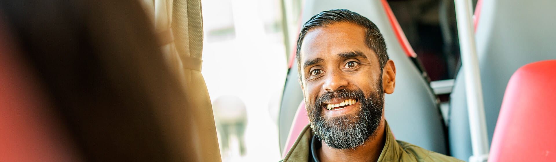 En man med silverstänkt skägg ler mot kameran och sin medpassagerare på bussen.